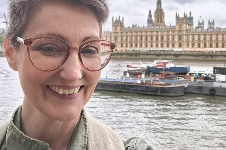 Bonnie has short cropped hair and is wearing glasses, smiling at the camera. She is stood opposite the Houses of Parliament in London 