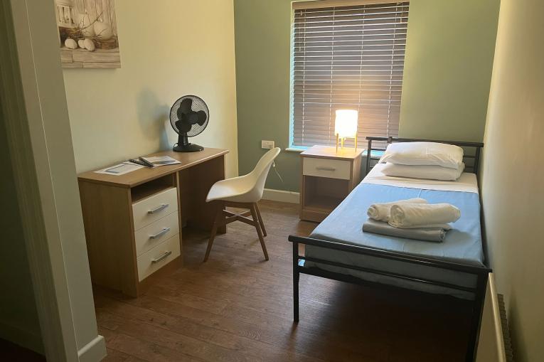 A single bed next to the wall with a desk, chair and shelf opposite