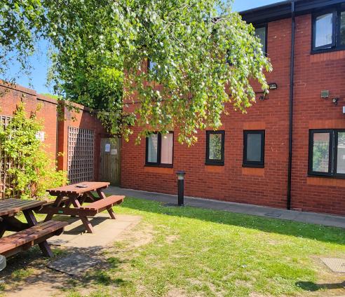 A garden. The Park House building is in the back of the shot with red brick walls. there is a brick wall along the left hand with trellis along the wall. There are two wooden picnic benches underneath a tree.  