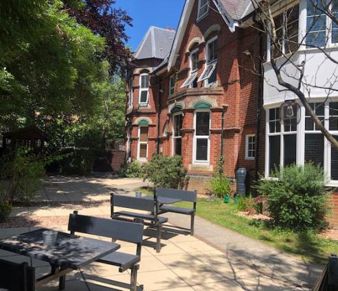 A photo of a garden in front of the red brick building. There is a patio with multiple tables and chairs and plenty of trees and bushes around the edge.