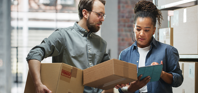 A man and a woman stand together looking at notepad in a brightly lit room. The man is holding cardboard boxes and there are many cardboard boxes stacked behind them in the room.