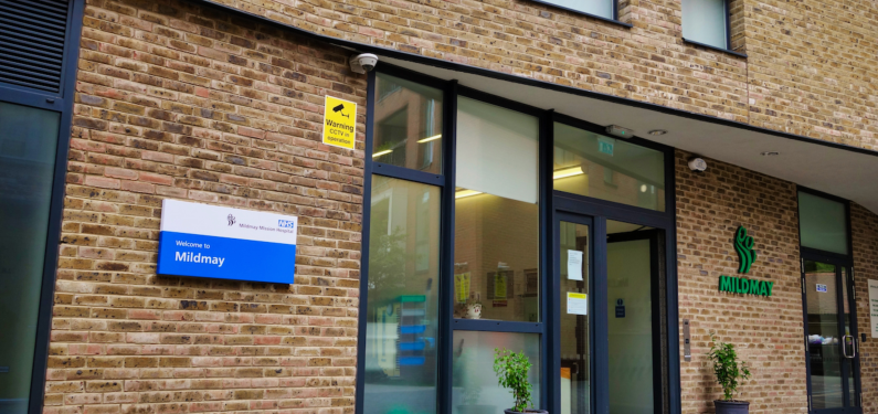 The front of a hospital with Brick wall and big glass double doors leading inside. A NHS sign is on the wall for Mildmay Hospital
