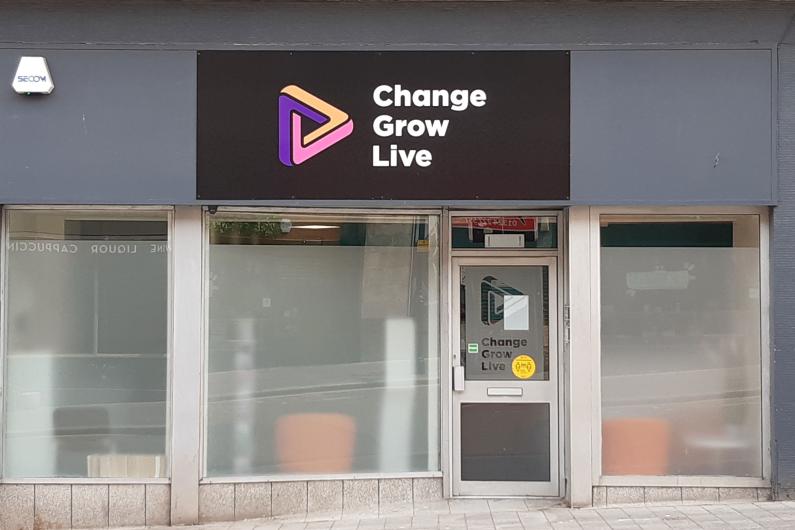 An image of the front of a building. There are two windows on the left side of the image, then a glass door, then another window on the right. There is a black sign above the doors and windows with a purple, orange and pink triangle with the words Change Grow Live written in white.