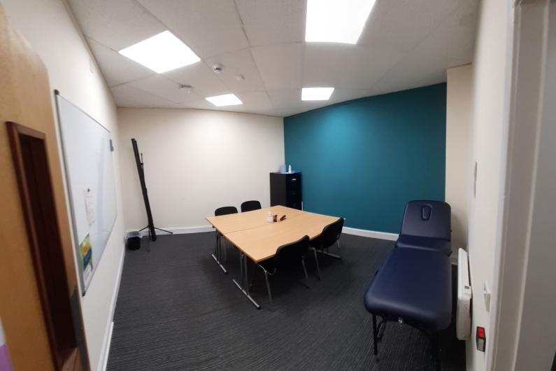 A photo of a room, there is a door in the bottom left hand corner which opens into the room, a blue wall on the right hand side of the image, the rest of the walls are white. There is a table in the middle of the room with four chairs around it.