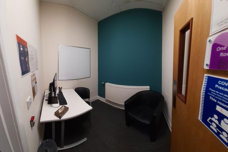 A photo of a room, there is a door on the right hand side of the photo, a blue wall next to the door and the rest of the walls are white. There is a table in the corner of the room and a couple of chairs.