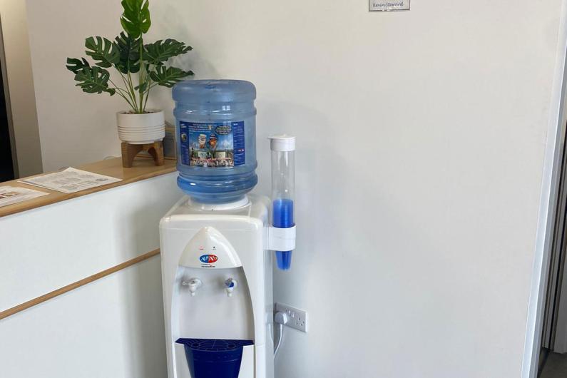 A photo of a water dispenser against a white wall and a reception desk.