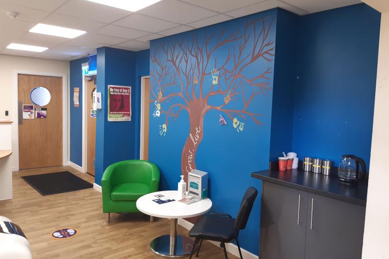 Macclesfield Recepton area - a room with a bright blue wall with a tree on it, green seating and white tables.
