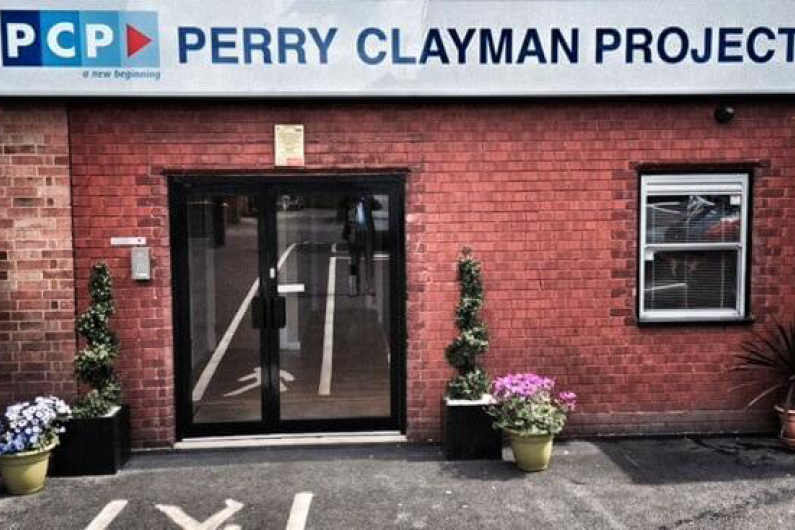 A red brick wall with a double glass door. A Perry Clayman Project sign hang above