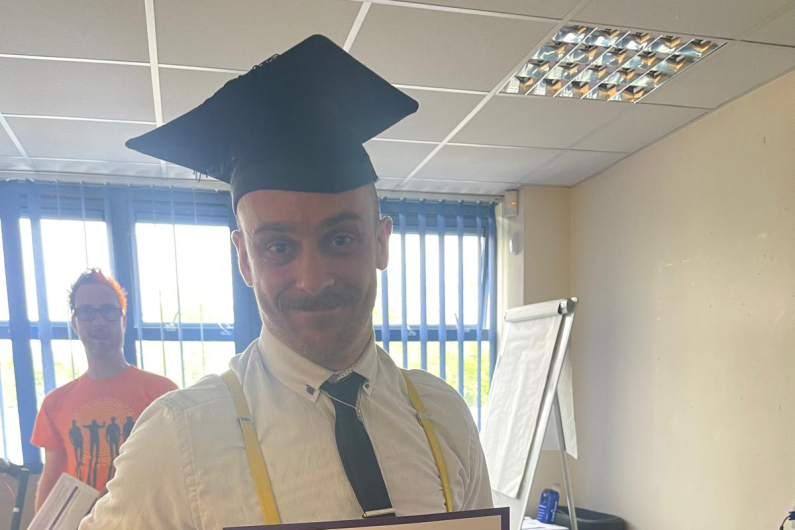 A man stood holding a certificate and wearing a graduation cap