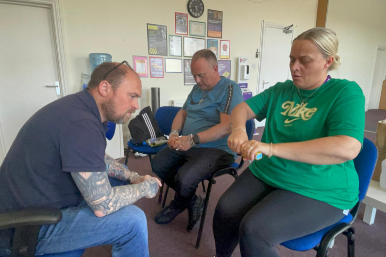 Three people sat on chairs, one is facing the other two. They are holding yellow Naloxone boxes