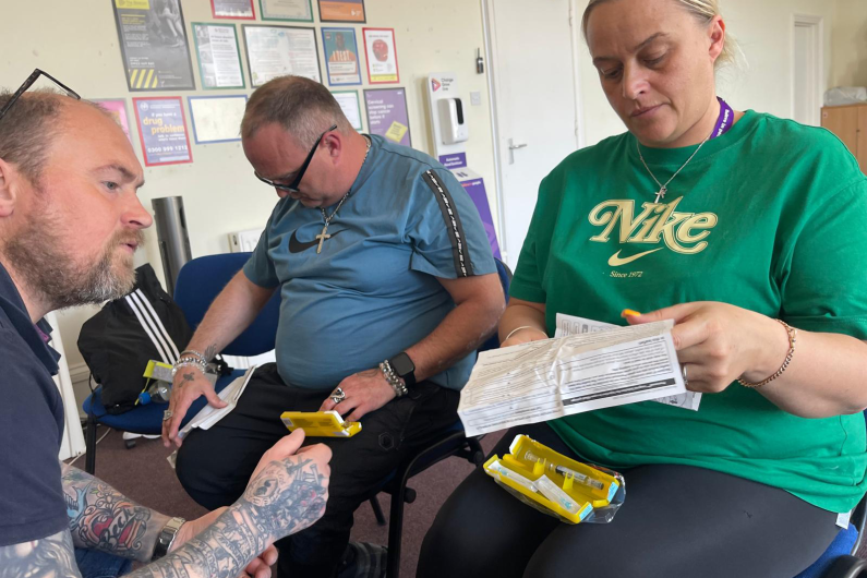 Three people sat on chairs, one is facing the other two. They are holding yellow Naloxone boxes