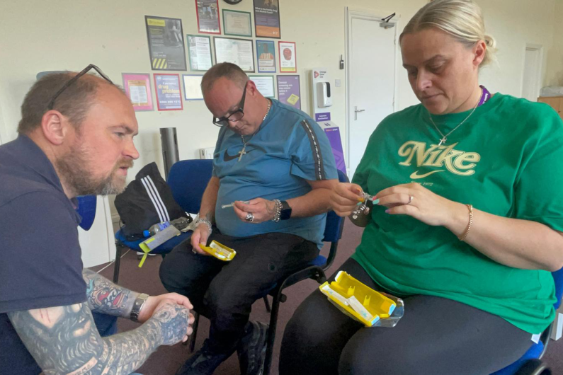 Three people sat on chairs, one is facing the other two. They are looking at yellow Naloxone boxes