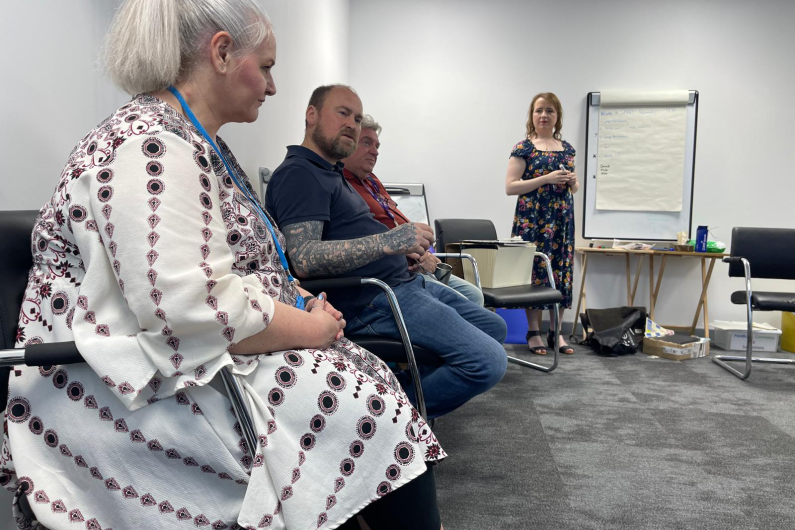 People sat in chairs around the edge of the room with a speaker stood in front of them