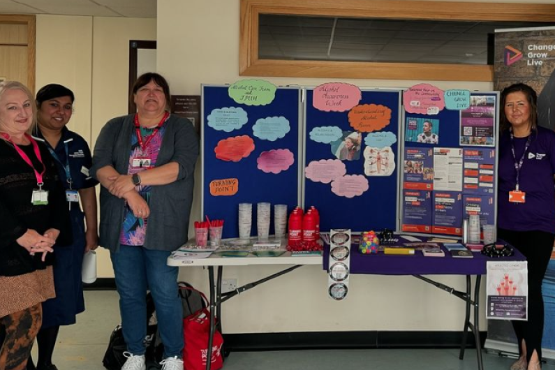 Four people stood either side of a table with a display board with posters and information around alcohol awareness week