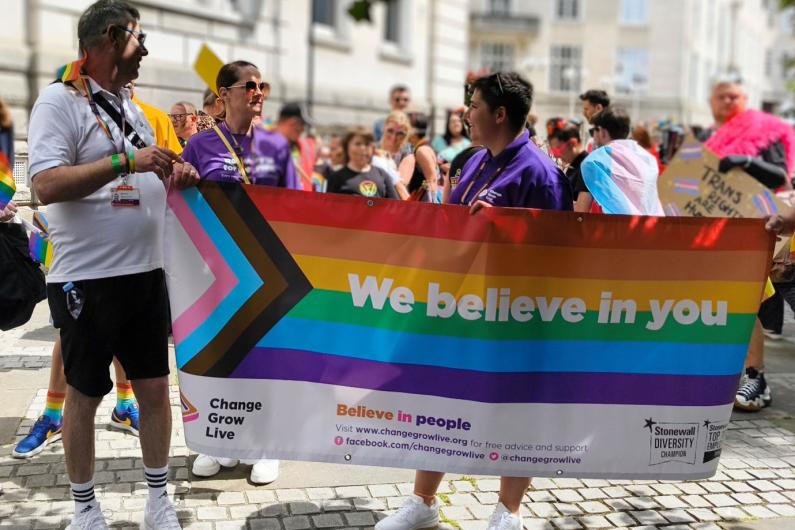 A group of people stood in the shade holding up a pride banner which reads "We believe in you"