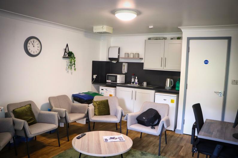 A room with a small kitchen area and chairs dotted around the room