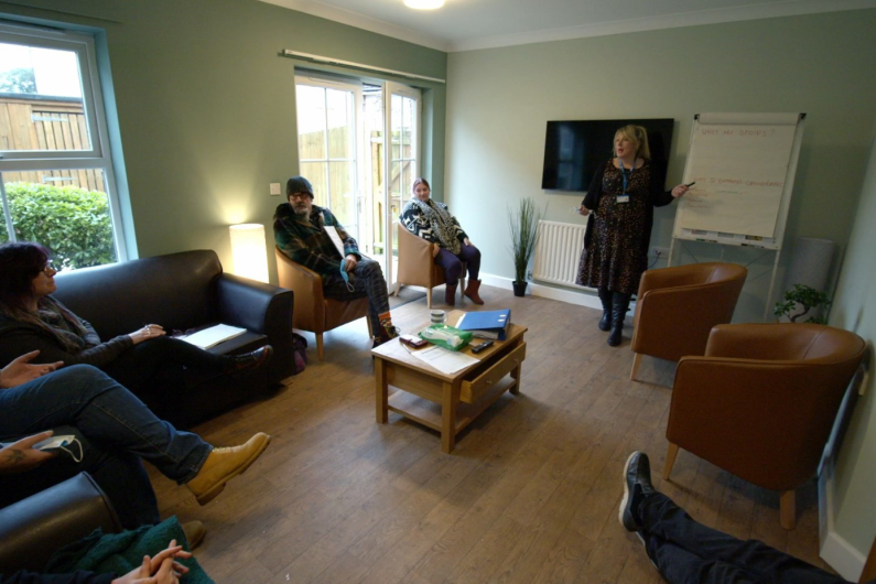 A room with leather sofas and chairs. A woman is stood at one end pointing at a flipchart while four other adults, are sat on the chairs looking at her. 