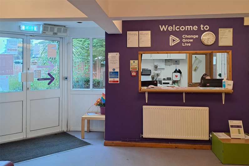 A reception area that has a hatch in the wall to speak to someone and a door leading outside.