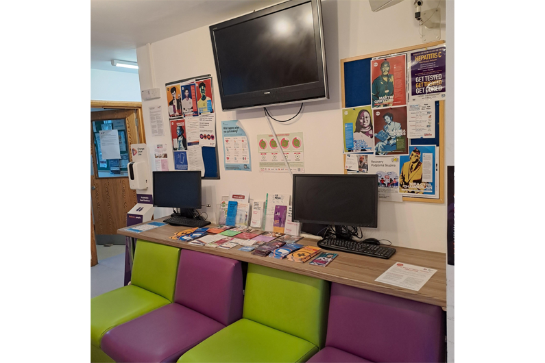 A line of alternating purple and green sofa chairs in front of a desk with two computers and a notice board